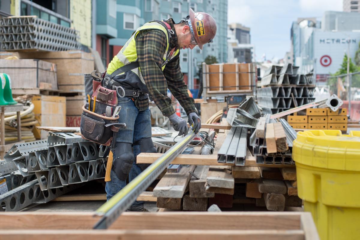 Construction worker measuring steel bracket on urban rooftop with city skyline and building materials in background.