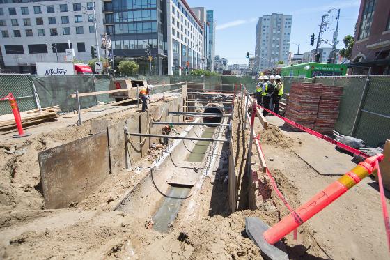Construction site showing underground excavation with steel framework, utility pipes, and workers in safety gear on urban street.