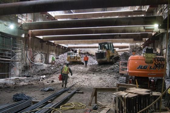 Construction work underway in underground transit station with heavy equipment, workers, and temporary steel supports.