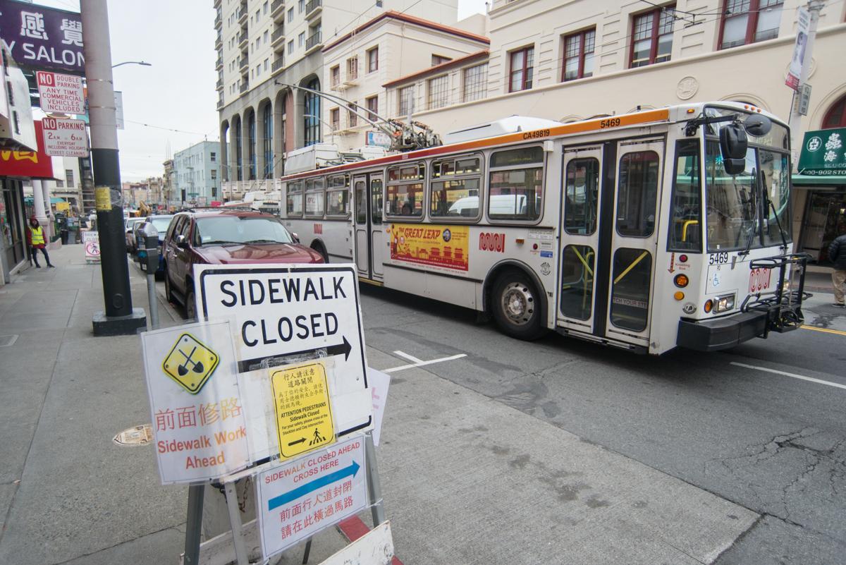 Sidewalk closed sign on urban street with bus and buildings in background.
