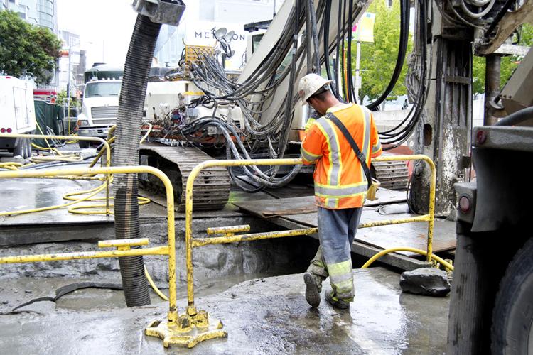 Worker in safety vest operating ground stabilization equipment on city street during construction.
