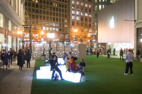 Evening urban plaza with illuminated benches, pedestrians, and downtown buildings.