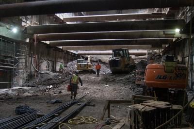 Construction workers and equipment operating inside an underground transit station headhouse with temporary steel supports and overhead lighting.