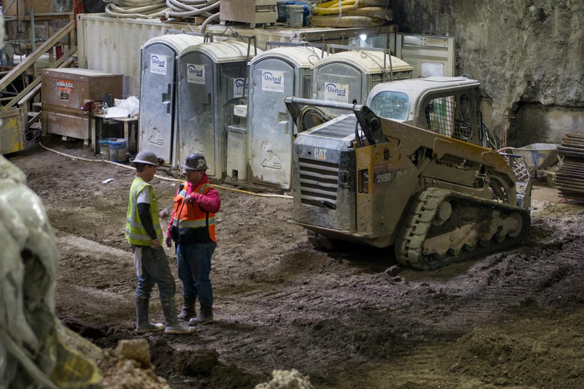 Two construction workers in safety gear stand near a CAT tracked loader at an underground transit construction site with portable trailers.