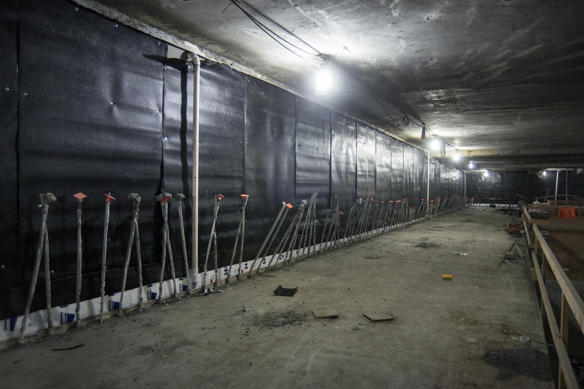 Underground transit station under construction with black waterproofing sheets lining concrete walls and support posts.