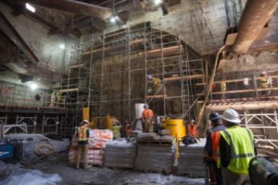 Construction workers in safety gear inside a reinforced underground excavation site with steel framework and equipment.