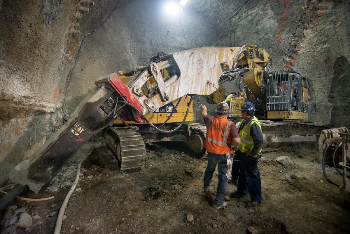 Excavator with jackhammer attachment breaking out rock in underground transit tunnel cavern.