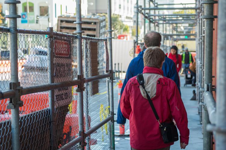 Pedestrians walking through covered transit corridor with chain-link fencing on both sides.