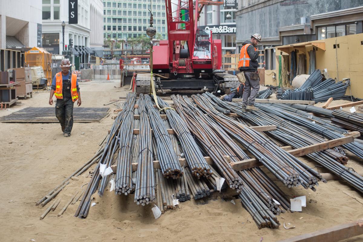 Construction workers organize steel rebar bundles at a downtown worksite with crane and equipment visible.
