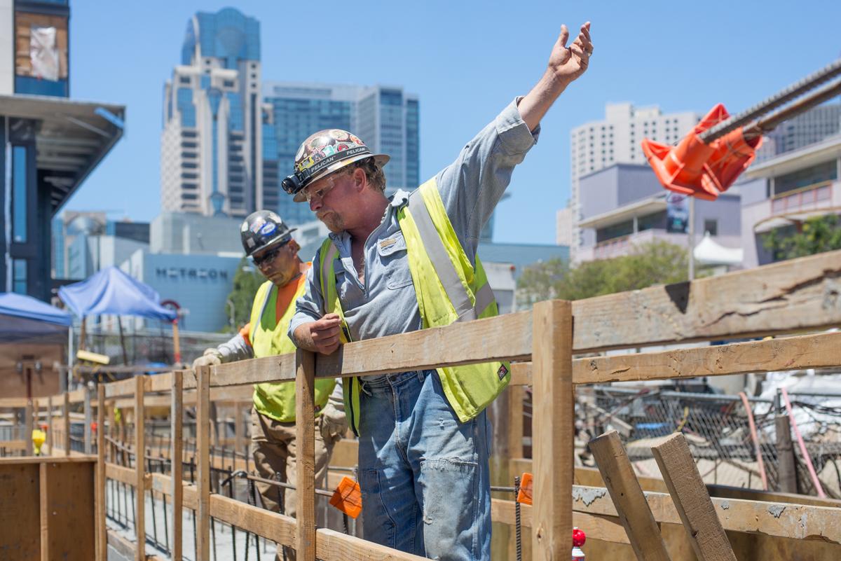 Construction supervisor in safety vest and hard hat directing crane operations on urban street with city skyline in background.