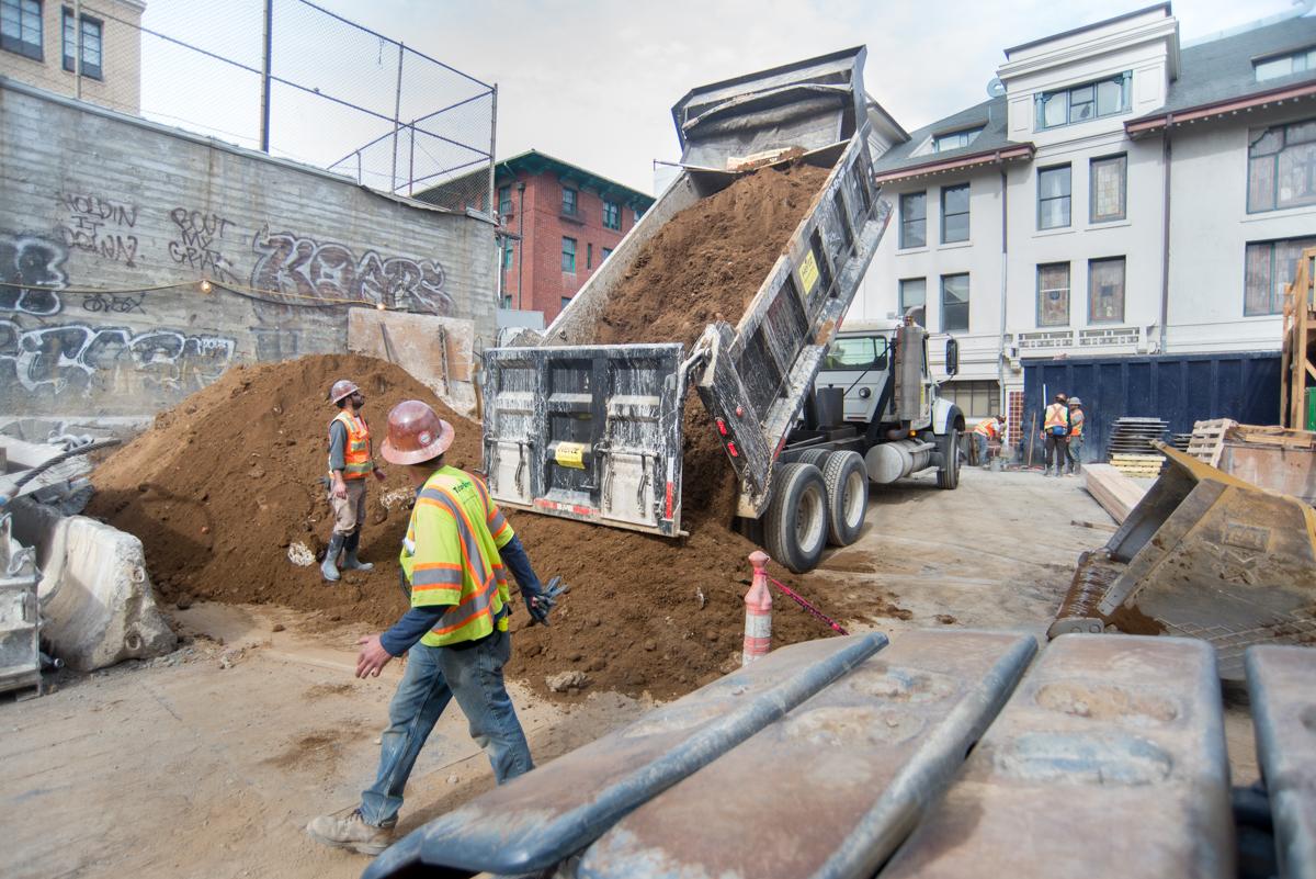 Construction workers guide dump truck unloading soil at urban transit construction site.