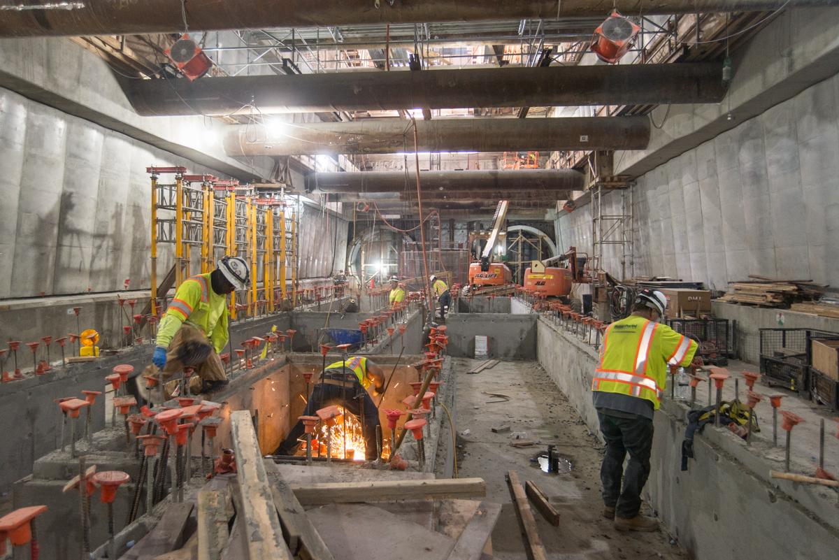 Construction workers in safety gear work on Union Square/Market Street Station platform during Central Subway tunnel construction.