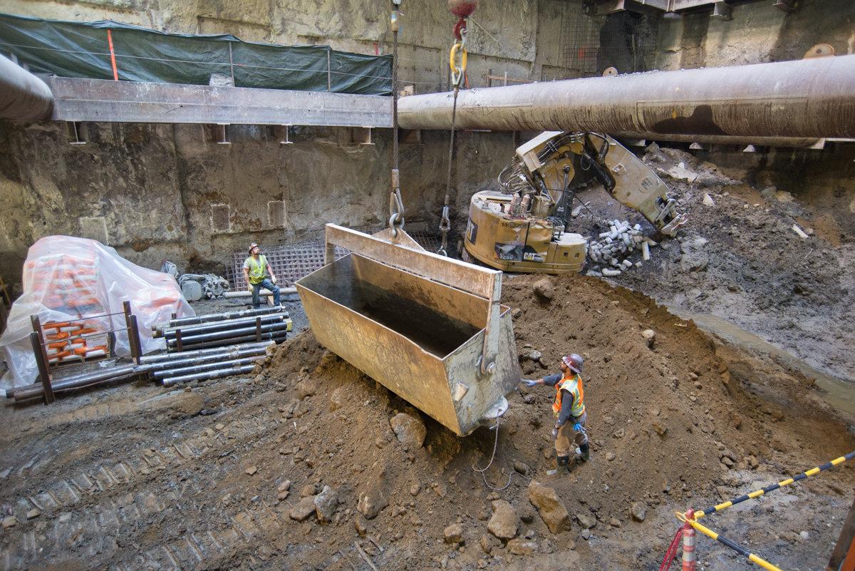 Workers guide a steel bucket suspended by crane cable in an underground construction shaft, with excavated material and equipment visible around the site.