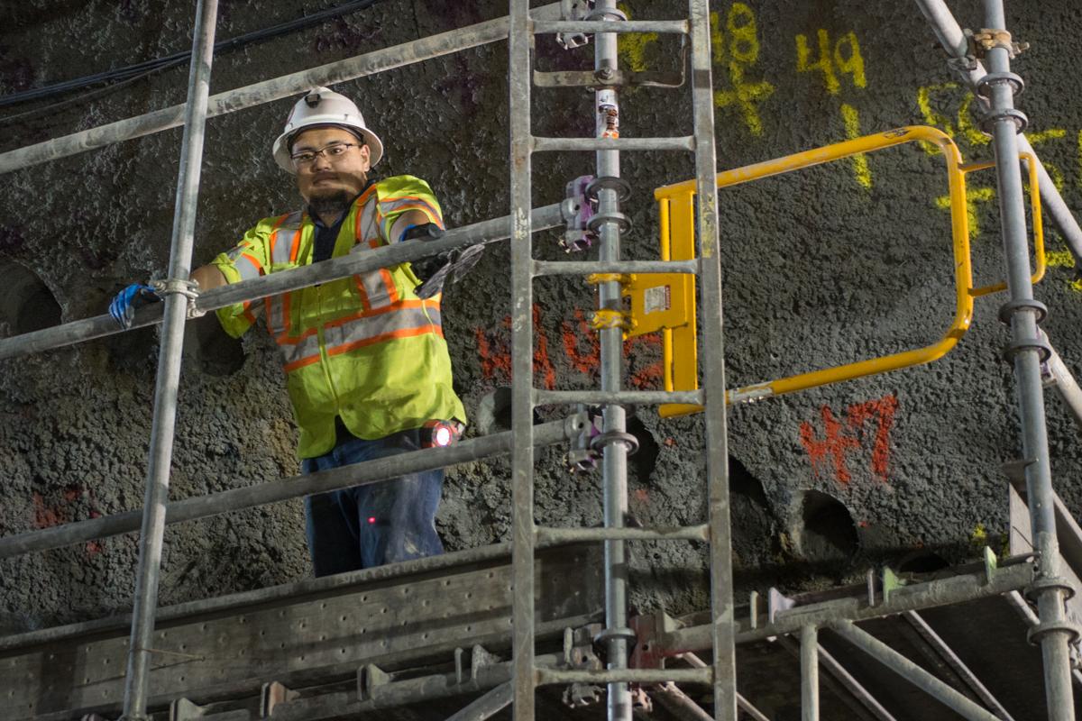 Worker in safety gear drilling pilot holes on scaffolding inside underground cavern for transit station construction.