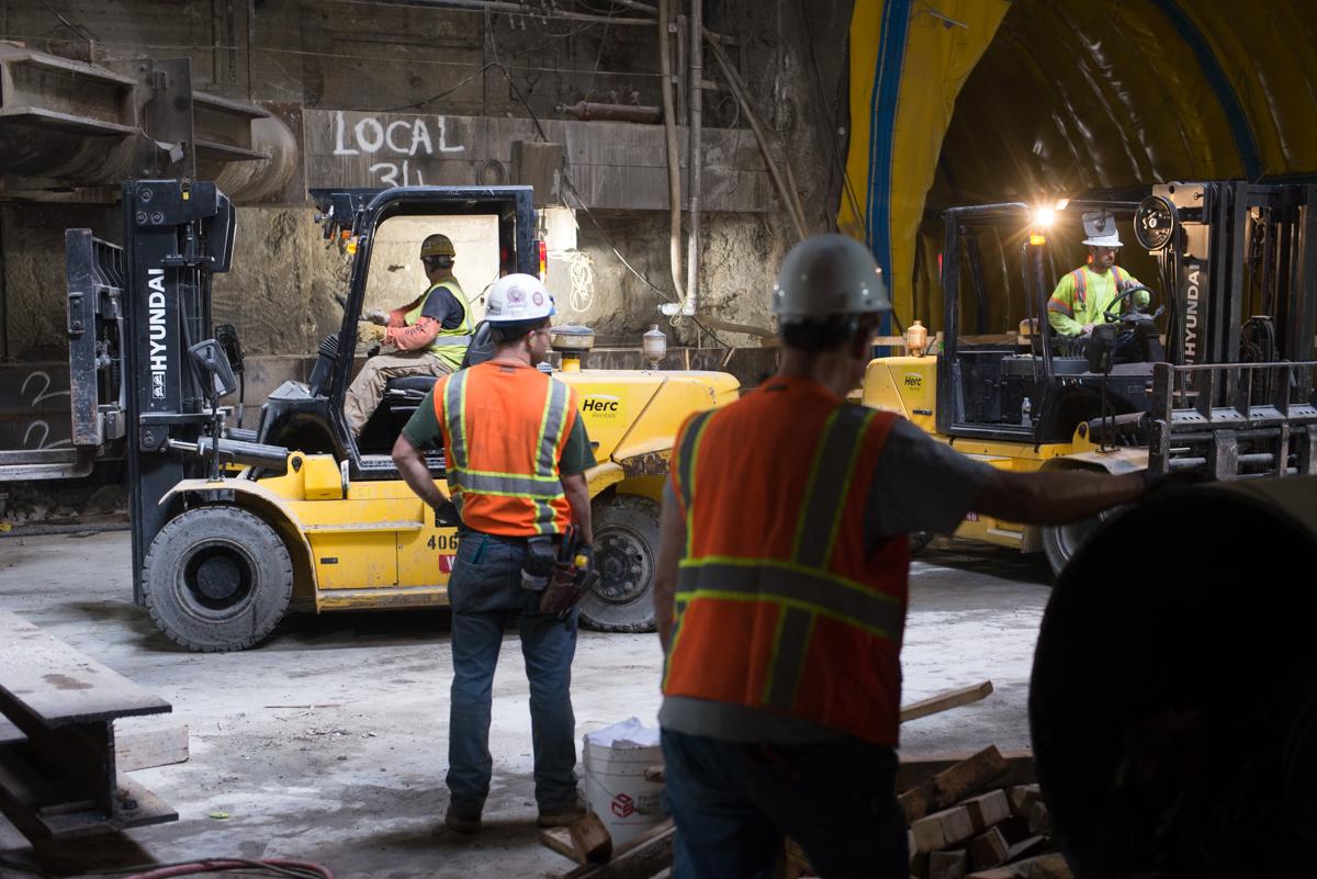 Workers guide forklift and crane operations while unloading supplies at underground transit station construction site.
