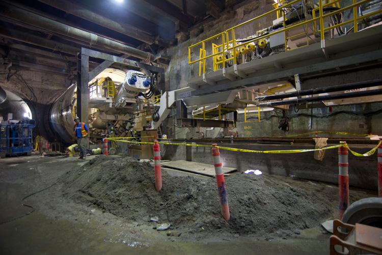 Workers prepare tunnel boring machine Big Alma in underground construction site with yellow safety railings and equipment.