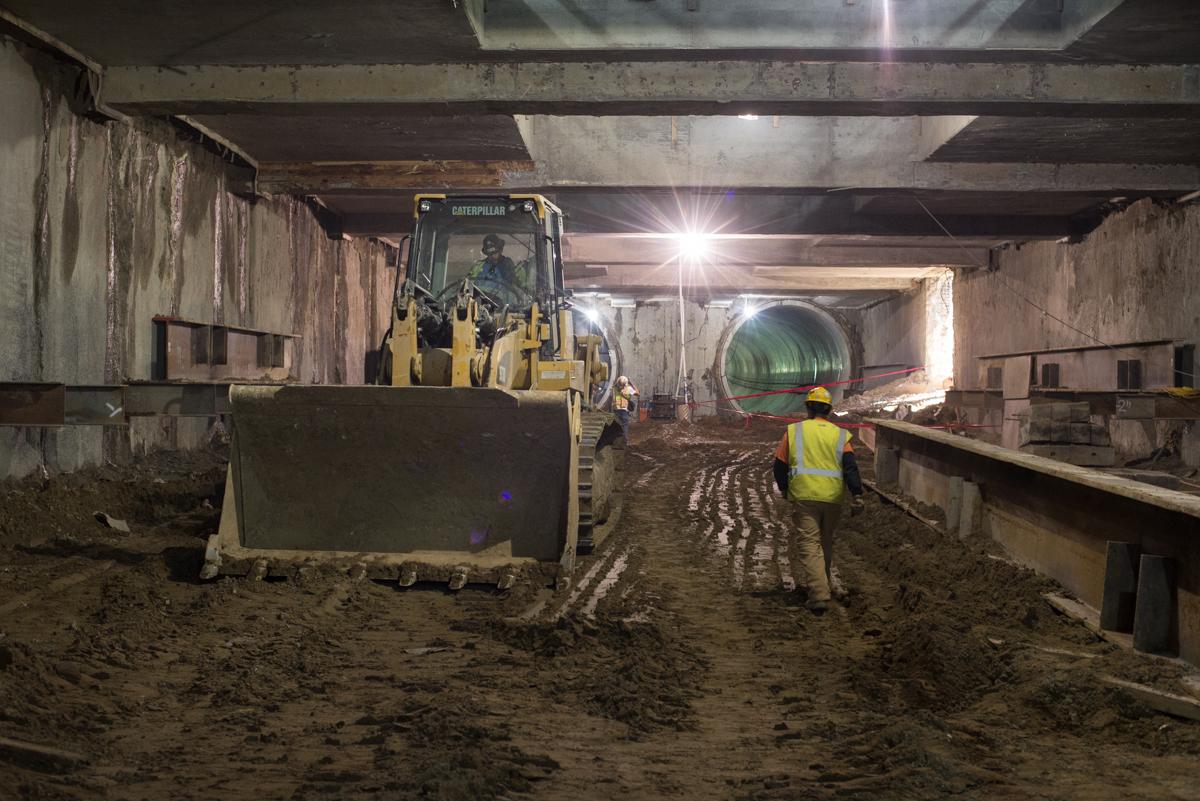 Construction worker in yellow safety vest observing Caterpillar loader moving soil inside underground transit station tunnel.