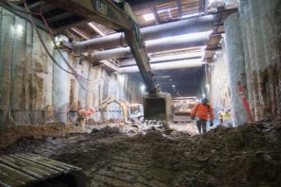 Workers in hard hats inside underground station box with exposed concrete walls and construction equipment.