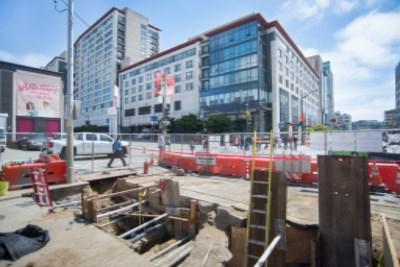 Construction excavation site on 4th Street with utility work barriers and urban buildings in background.