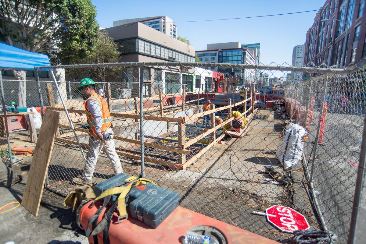 Construction site with fenced work area, wooden structures, and workers installing underground utilities on city street.
