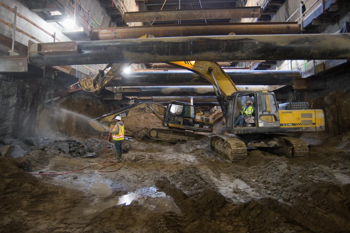Workers operate excavation equipment to remove concrete from station box walls underground.
