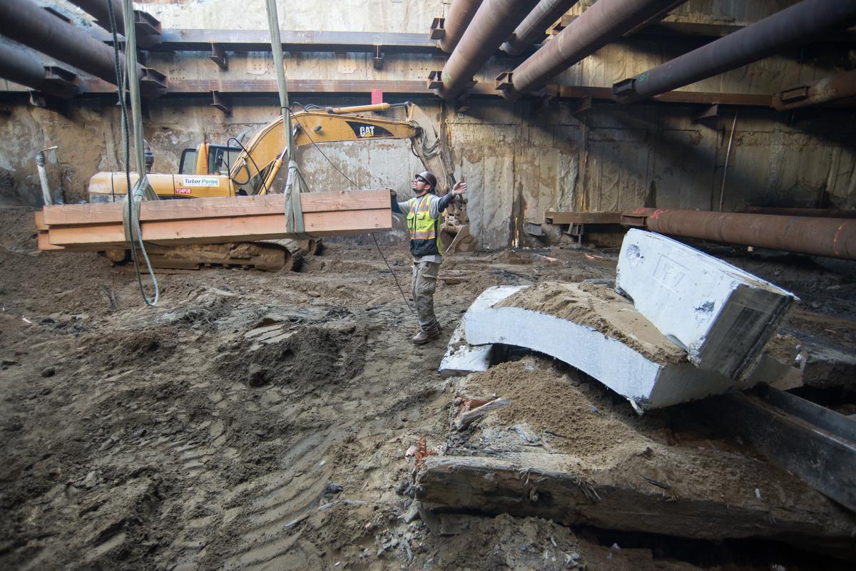 Construction worker directs excavator lowering steel beam into underground Central Subway tunnel.