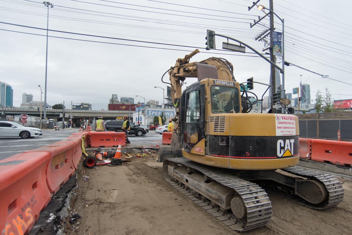 Caterpillar excavator operating in street work zone with orange barriers and construction equipment visible.