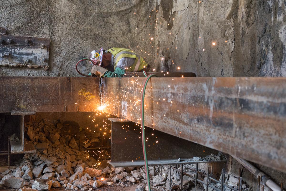 Worker in yellow helmet welding steel beam with sparks flying at construction site.