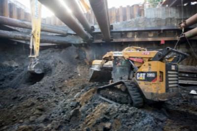 CAT excavator removing materials from underground station construction trench with large pipes overhead.