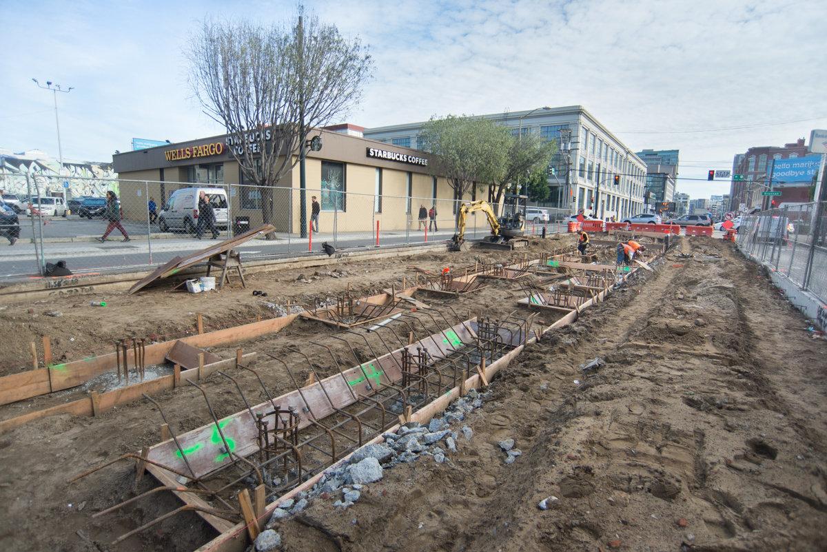 Construction site with exposed foundation work, rebar, and concrete forms along urban street with Starbucks and Wells Fargo visible.
