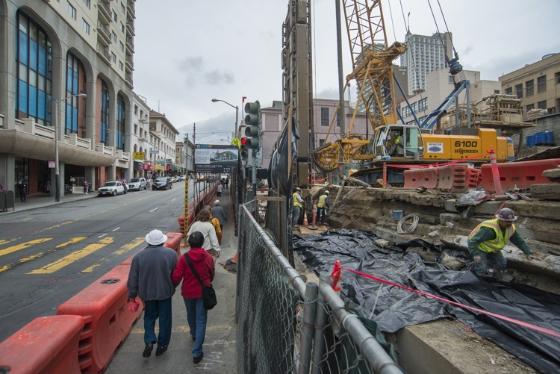 Construction site with hydromill equipment digging station walls on urban street with pedestrians and barriers.