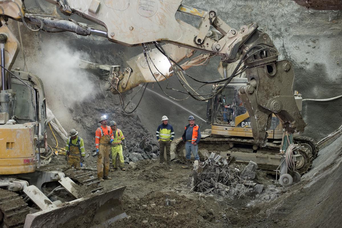 Workers in safety gear excavate underground station cavern with heavy equipment and machinery.
