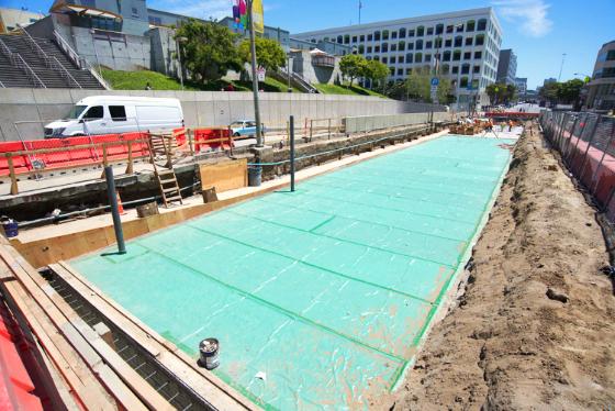 Construction site showing turquoise waterproofing membrane laid on underground station roof deck with surrounding excavation and barriers.
