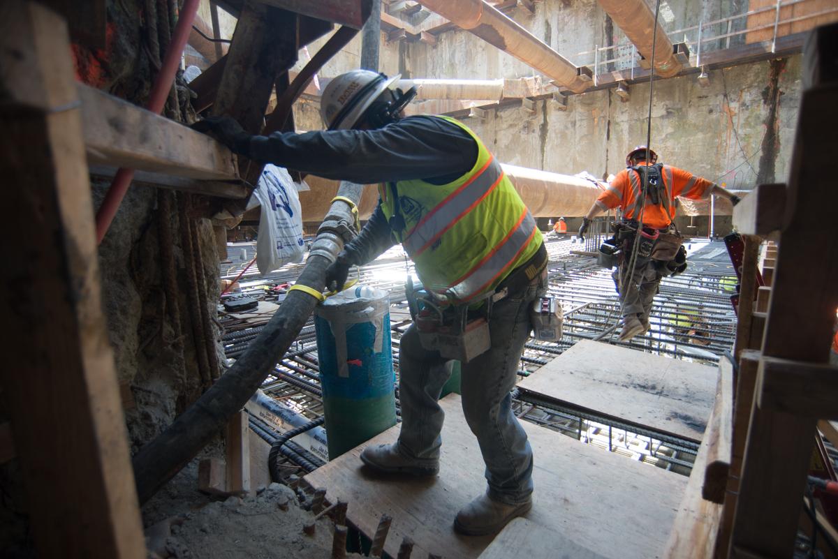 Construction worker in safety vest operating concrete pump hose at underground transit project site.