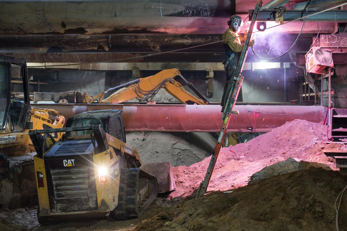 Underground construction site with excavator, CAT skid loader, and welder assembling steel truss under industrial lighting.