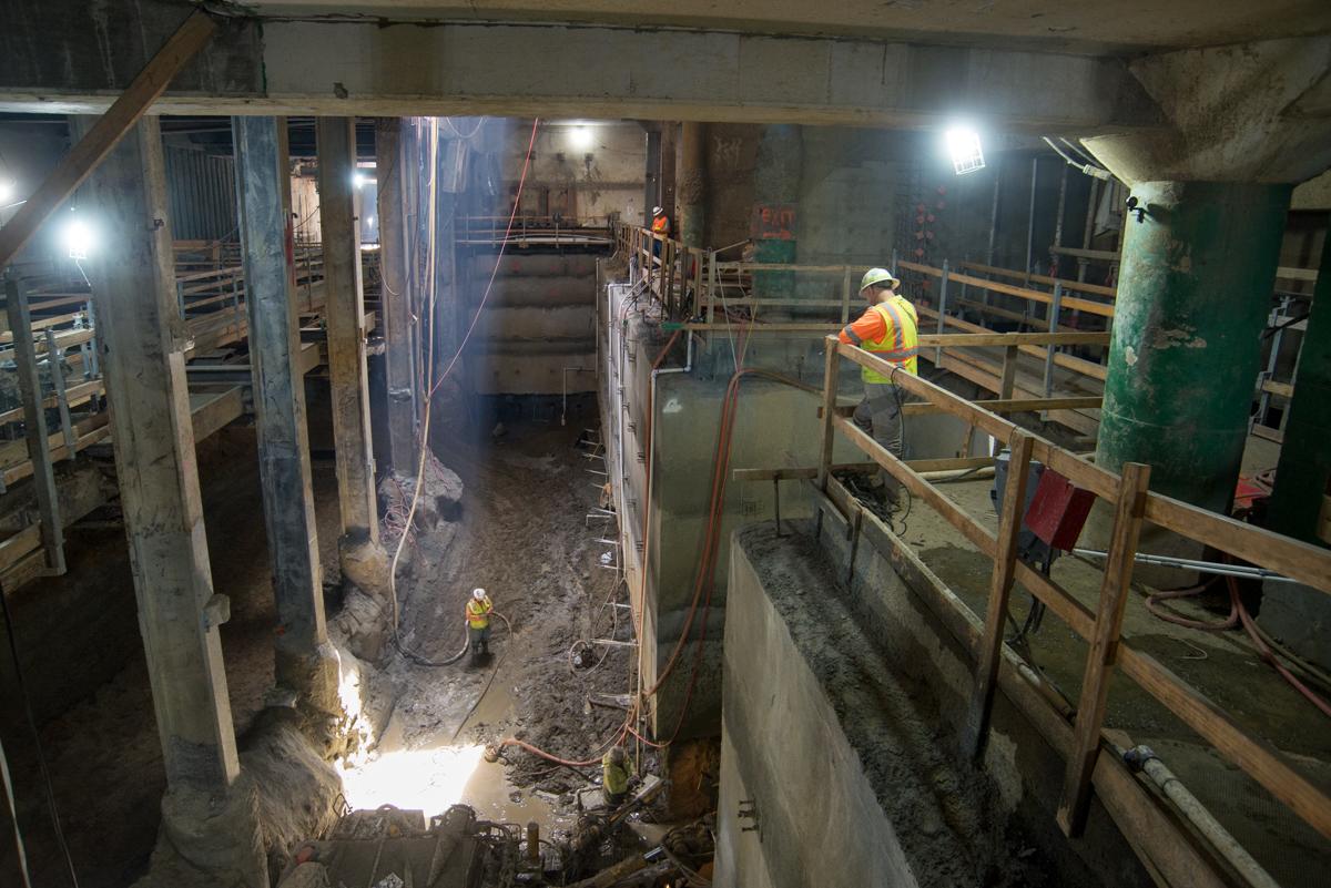 Workers in safety gear excavate underground station area with structural steel bracing and concrete supports visible.