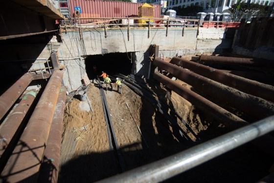 Underground construction site with workers positioning rebar in excavated portal area surrounded by safety barriers.