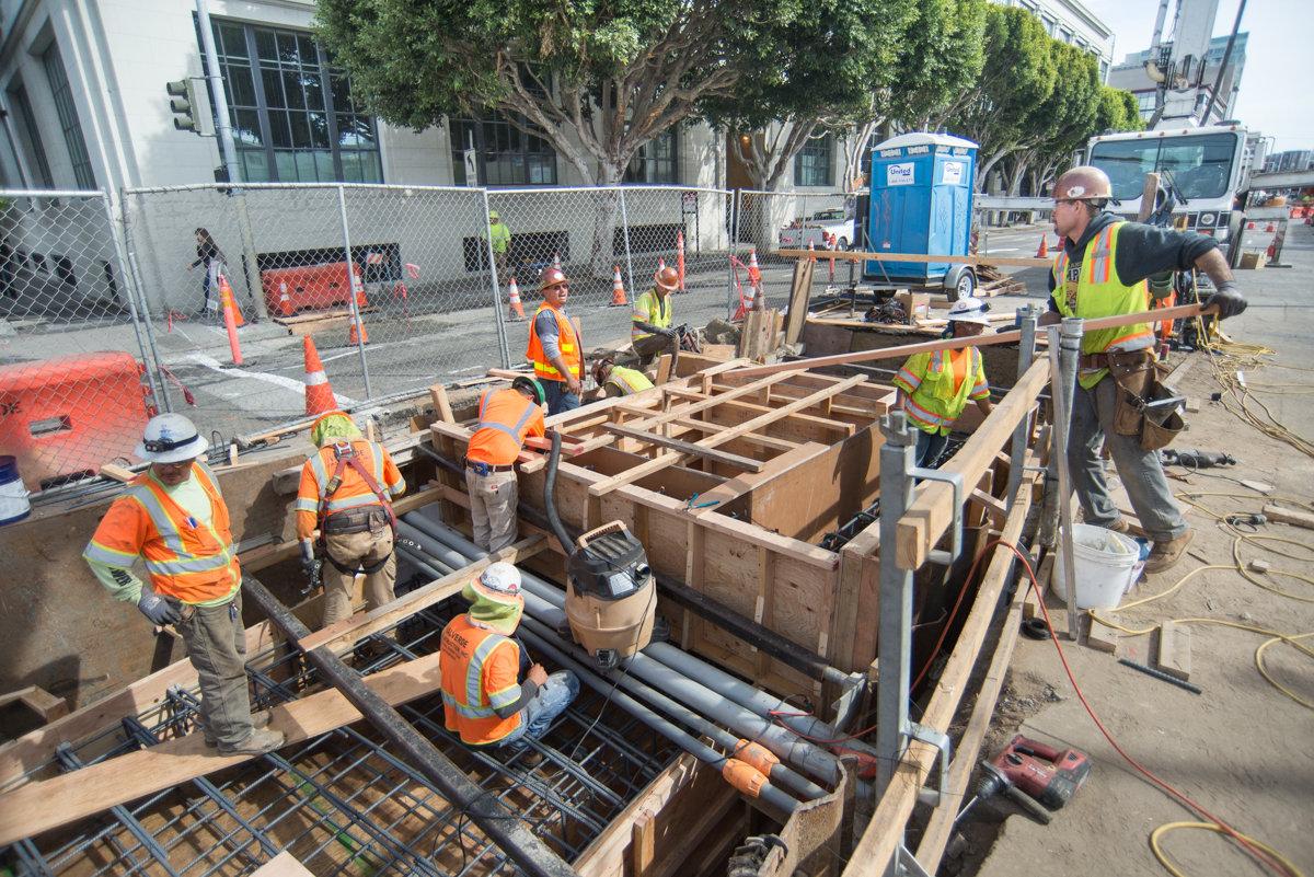Construction workers in safety vests assemble rebar and concrete forms at a subway construction site.