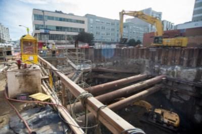 Construction excavation site with steel sheet piling, heavy equipment, and urban buildings in background.