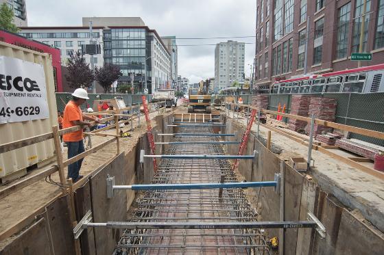 Construction site showing underground transit tunnel with steel reinforcement bars and wooden forms, urban street in background.