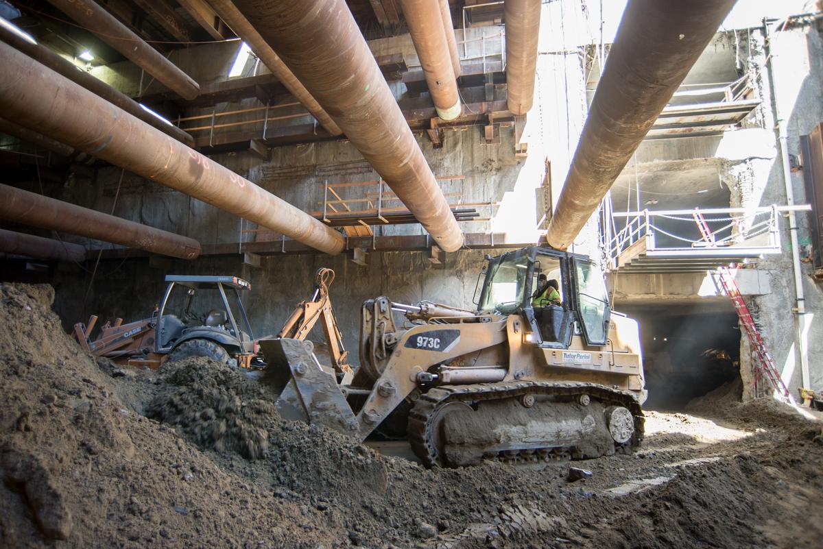 Bulldozers excavate soil beneath large concrete pipes at an underground transit station construction site.