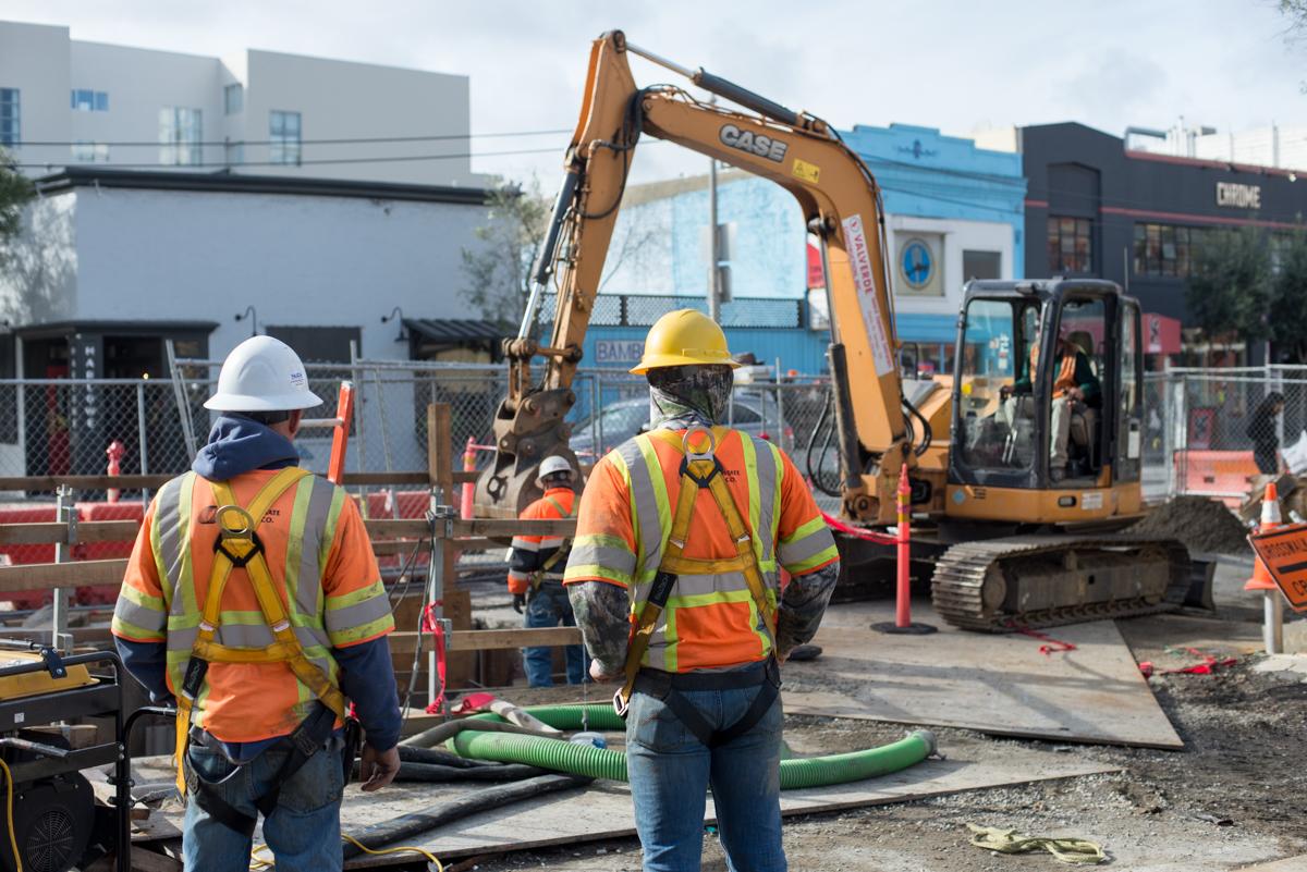 Construction workers in safety gear operate excavation equipment during street utility work.