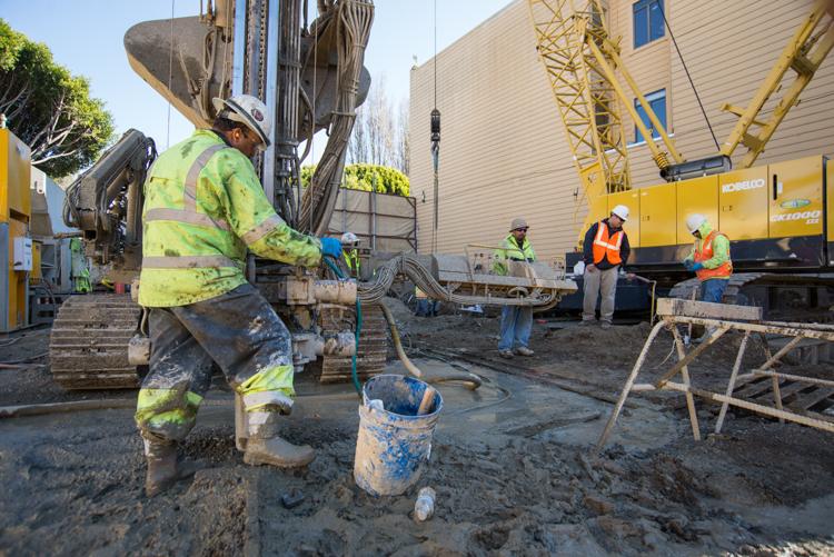 Construction workers in safety vests operate concrete pump truck at underground transit project site with yellow crane equipment visible.