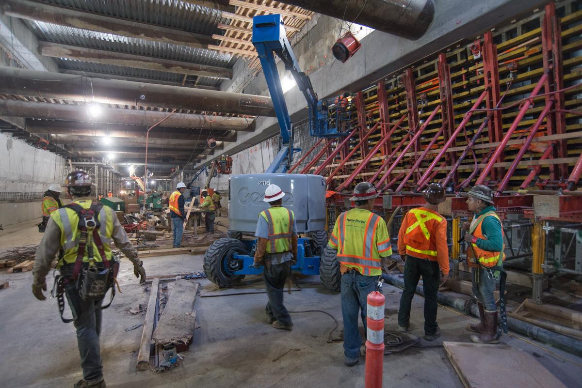 Construction workers in safety gear operate a blue boom lift near red structural forms inside an underground transit station.