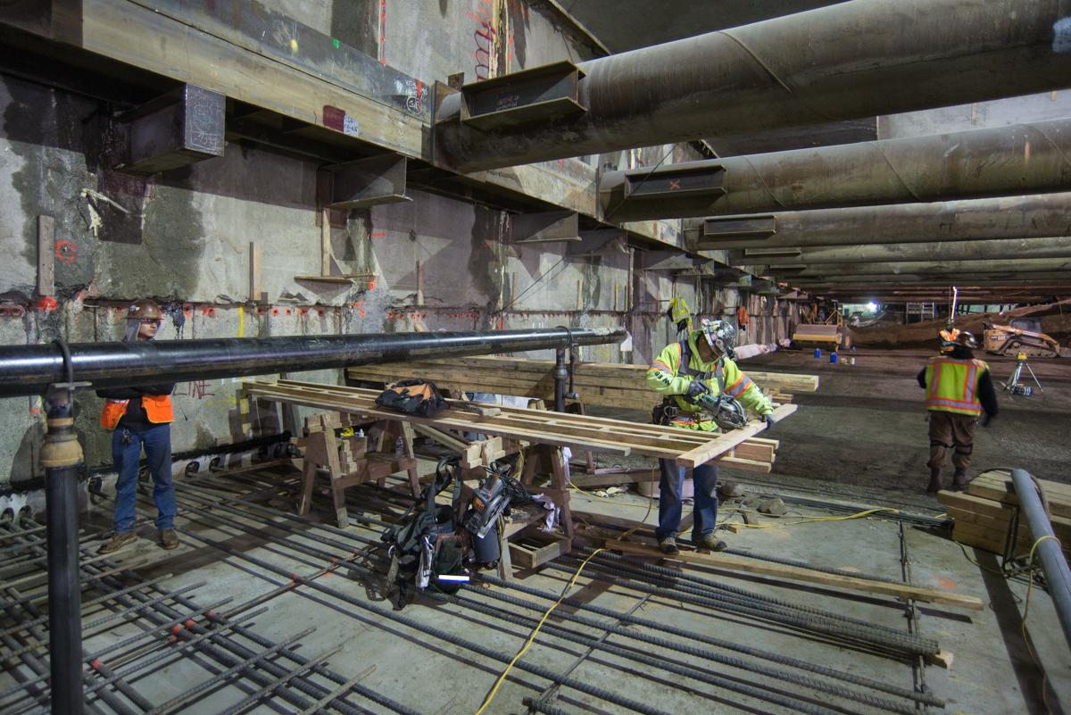 Workers in safety vests install rebar and construct concrete forms beneath elevated highway structure at transit station.
