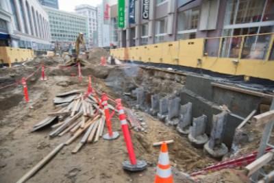 Construction site with exposed foundation, steel pilings, yellow equipment, and red safety barriers along urban street.