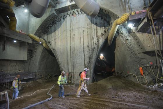 Workers in safety vests inside large tunnel excavation with concrete walls and yellow ventilation ducts.