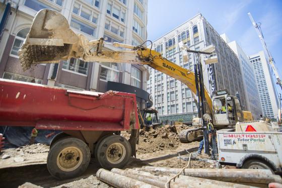 Construction equipment and dump truck at urban street excavation site with buildings in background.