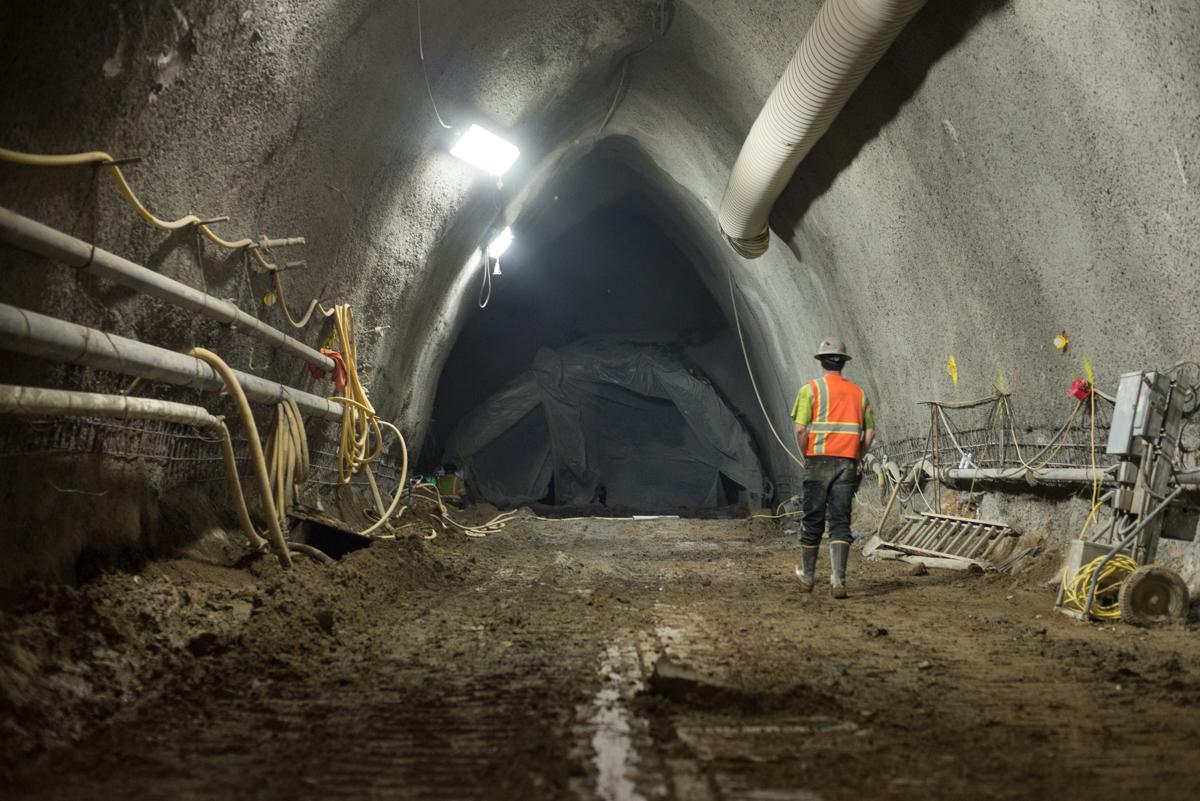 Worker in safety vest inspecting tunnel excavation with construction equipment and lighting.