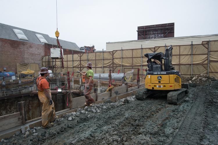 Construction crew operates yellow excavator at urban demolition site with brick buildings and safety barriers.